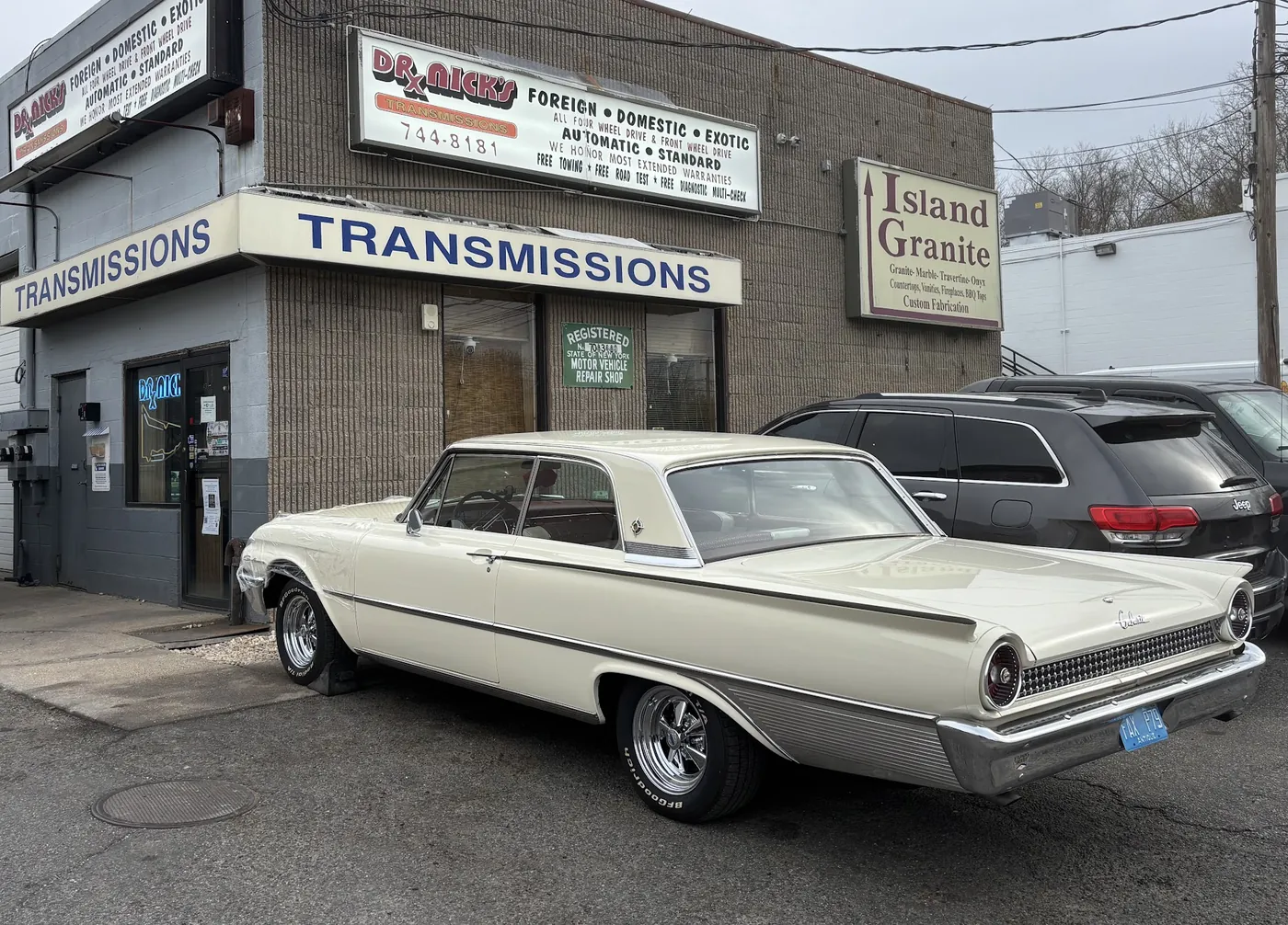 A white classic Ford Galaxie parked in front of the Dr. Nick's Transmissions storefront
